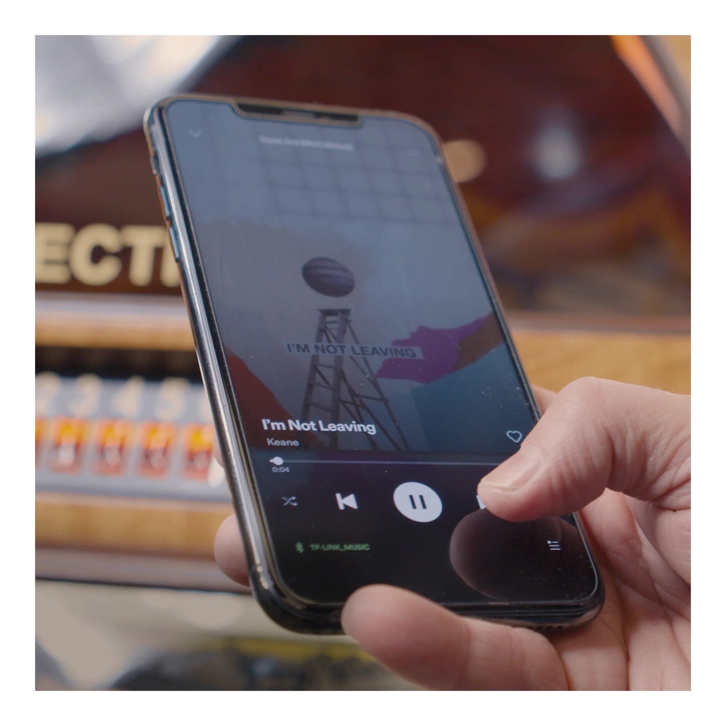 Hand holding a smartphone with a music app displayed, set against a blurred indoor background.