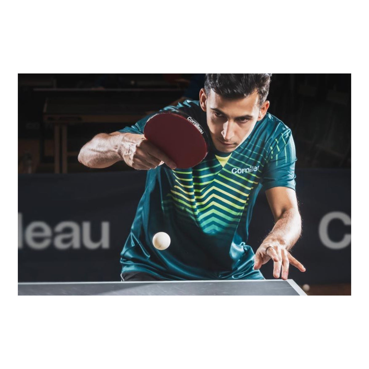 A man playing table tennis, wearing a teal and green striped athletic jersey with a red paddle, focused on a white ball in motion over a table tennis table.