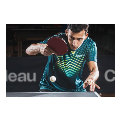 A man playing table tennis, wearing a teal and green striped athletic jersey with a red paddle, focused on a white ball in motion over a table tennis table.