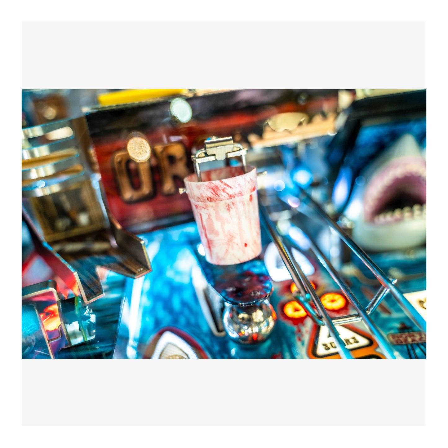 Close-up of a pinball machine with a glass of pink liquid on the playfield.