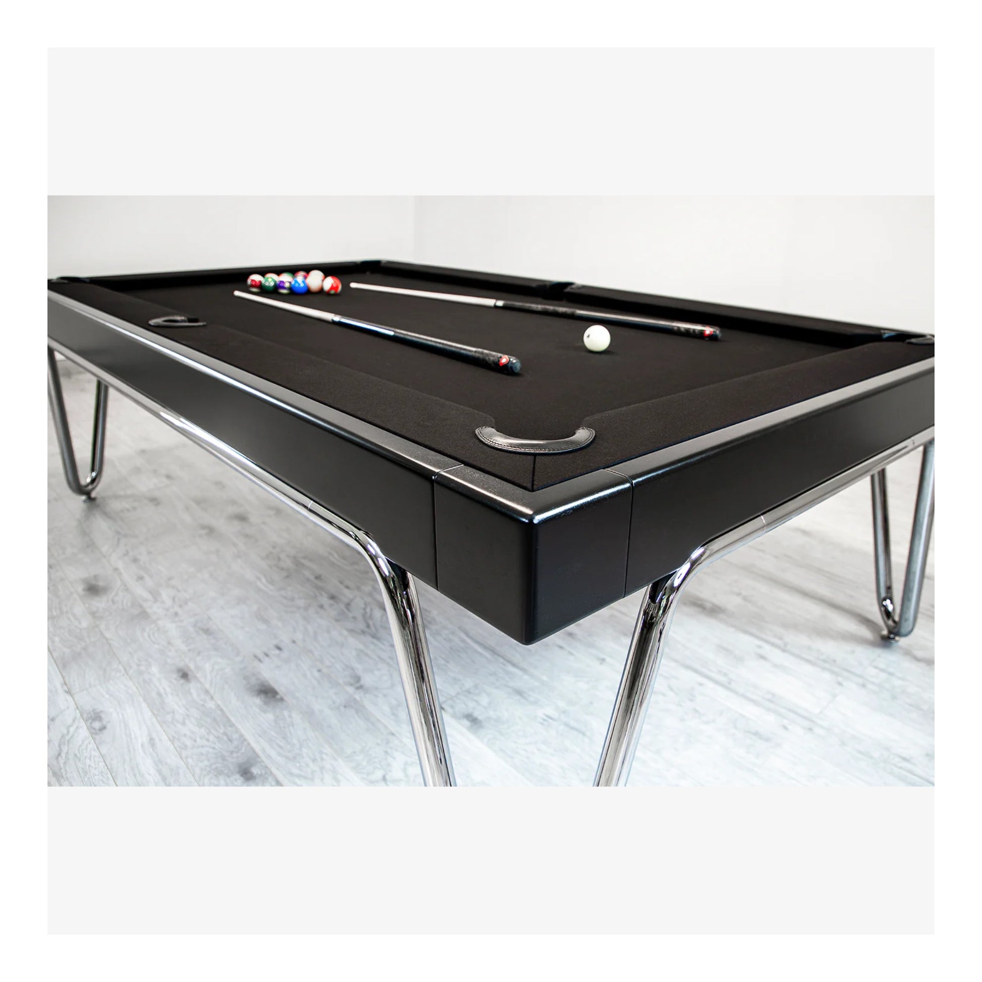 Black pool table with chrome legs on a light wooden floor.
