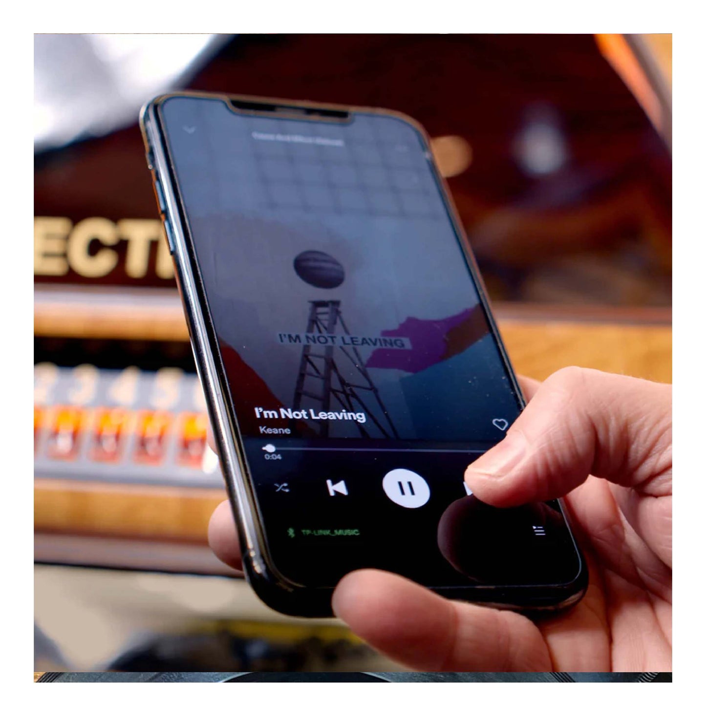 Hand holding a smartphone with a music app playing 'I'm Not Leaving' by Keane, with a guitar in the background.