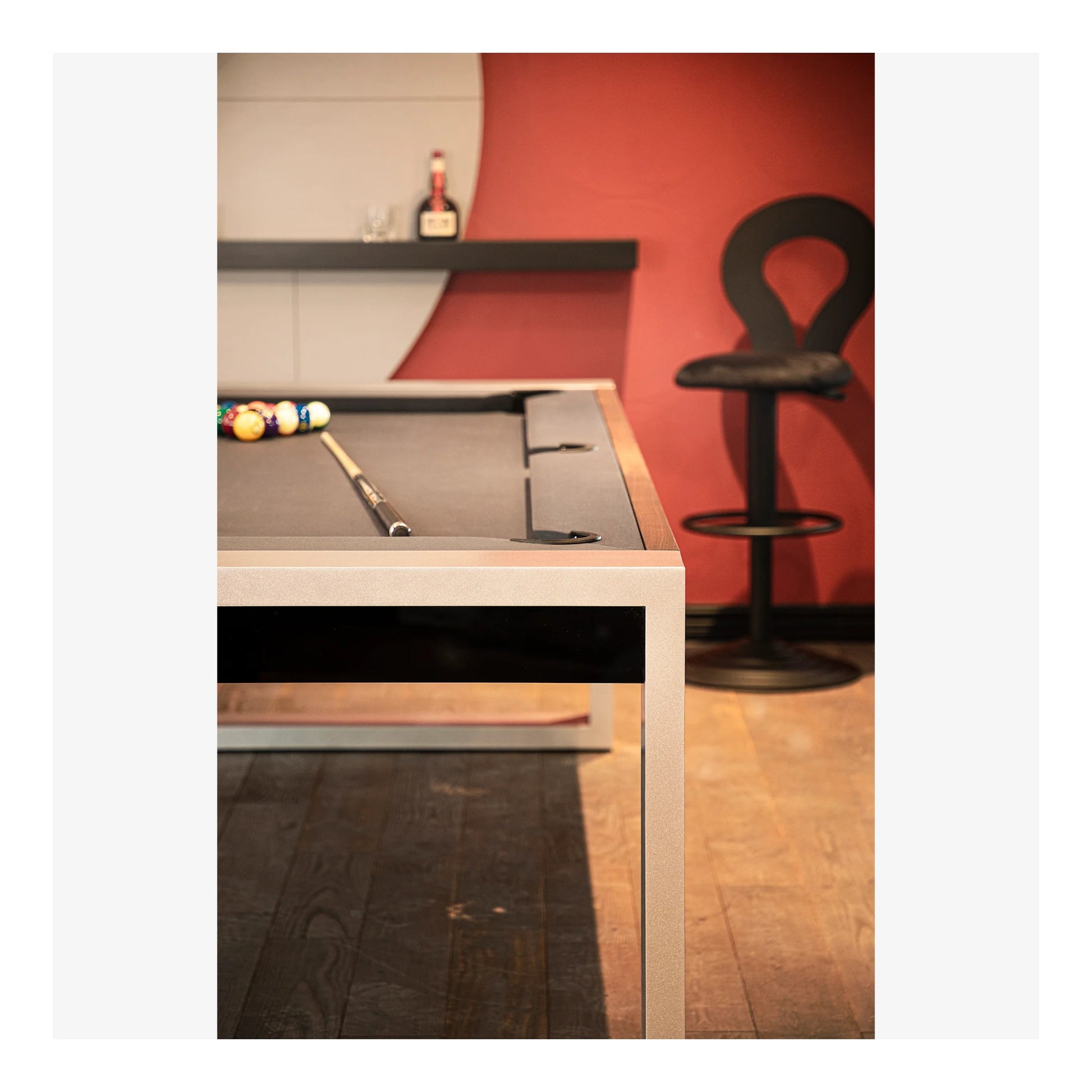 Pool table with cues and balls in a room with a red wall and bar stool.